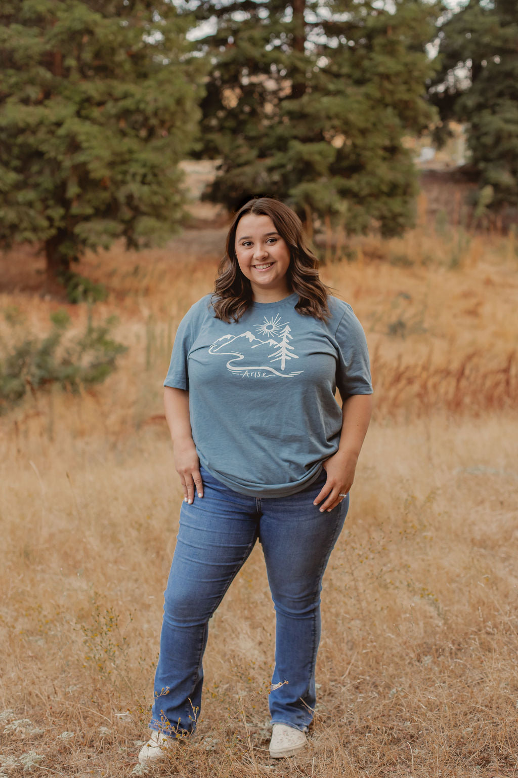 Woman wearing Arise T-Shirt in blue. Arise logo included white tree, mountain and sun