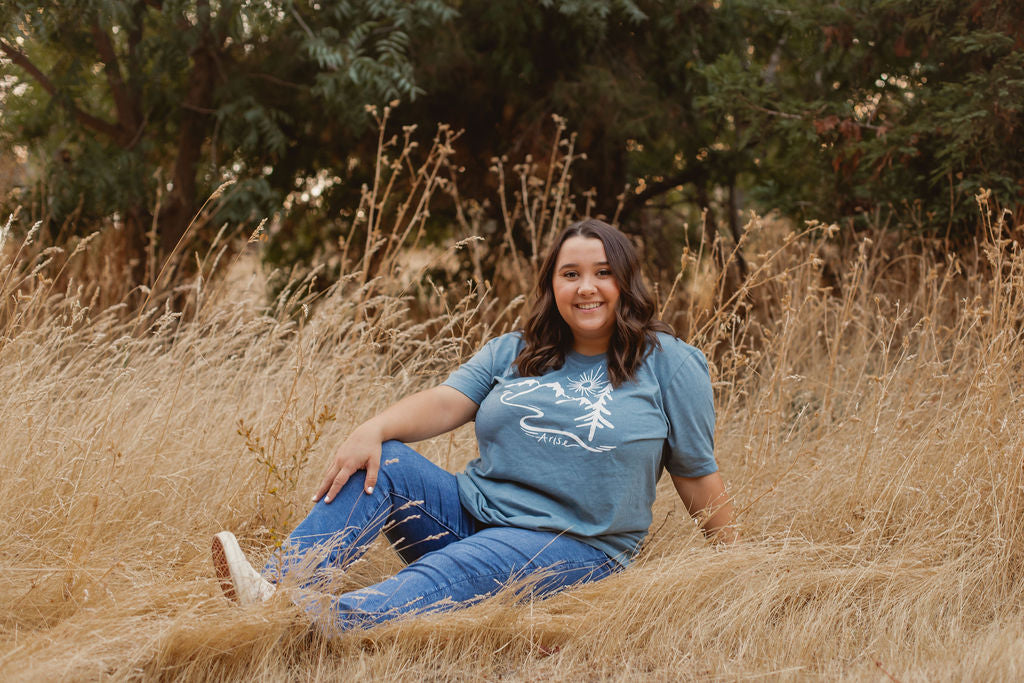 Woman wearing Arise T-Shirt in blue. Arise logo included white tree, mountain and sun