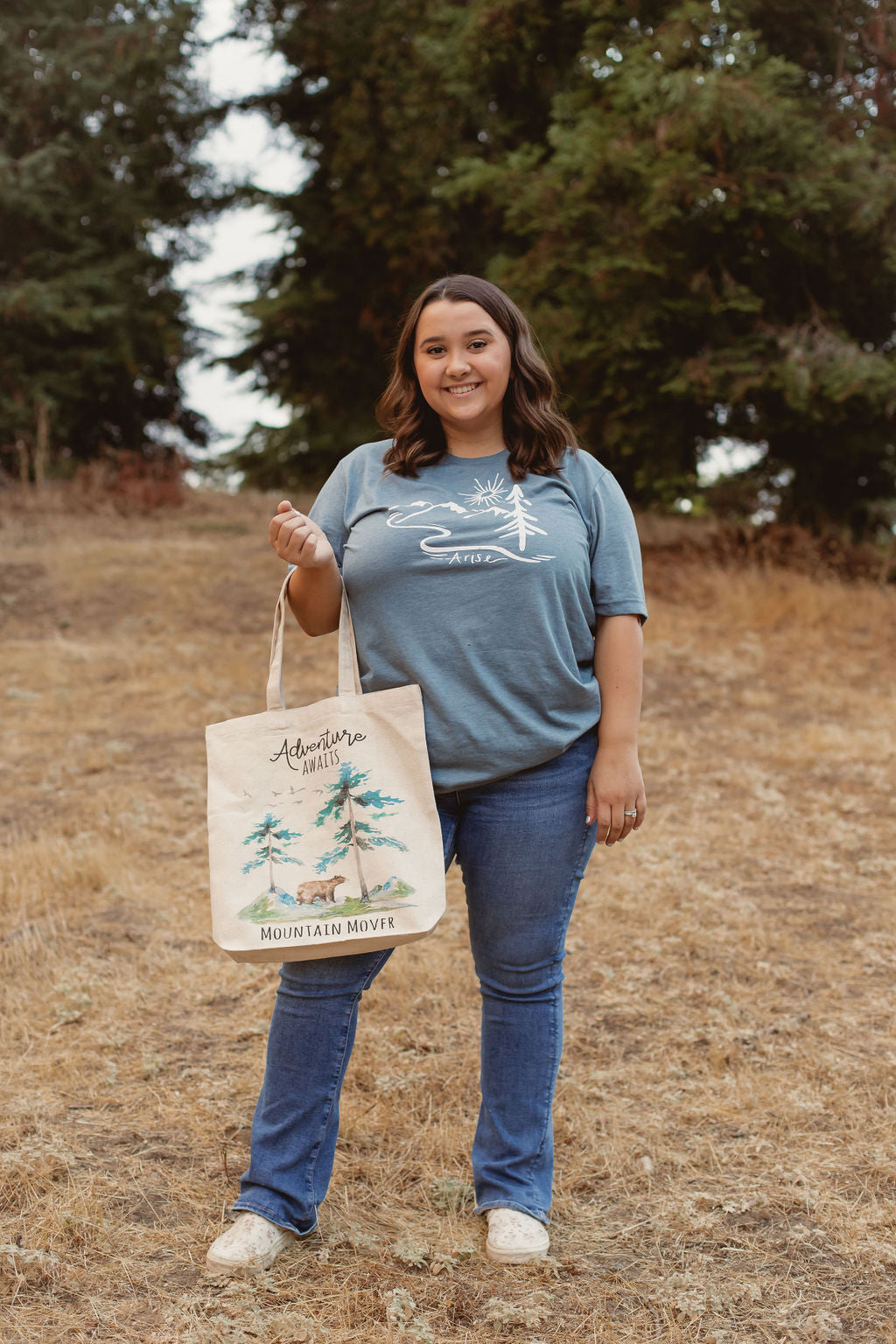 Woman in arise tee with white canvas bag that says adventure awaits with bear and mountain scene