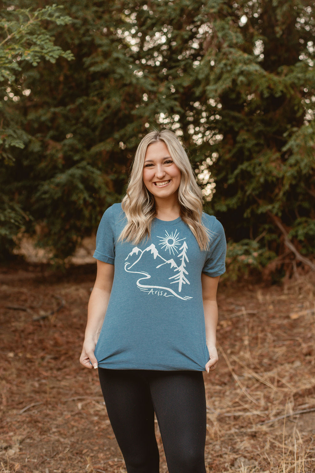 Woman wearing Arise T-Shirt in blue. Arise logo included white tree, mountain and sun
