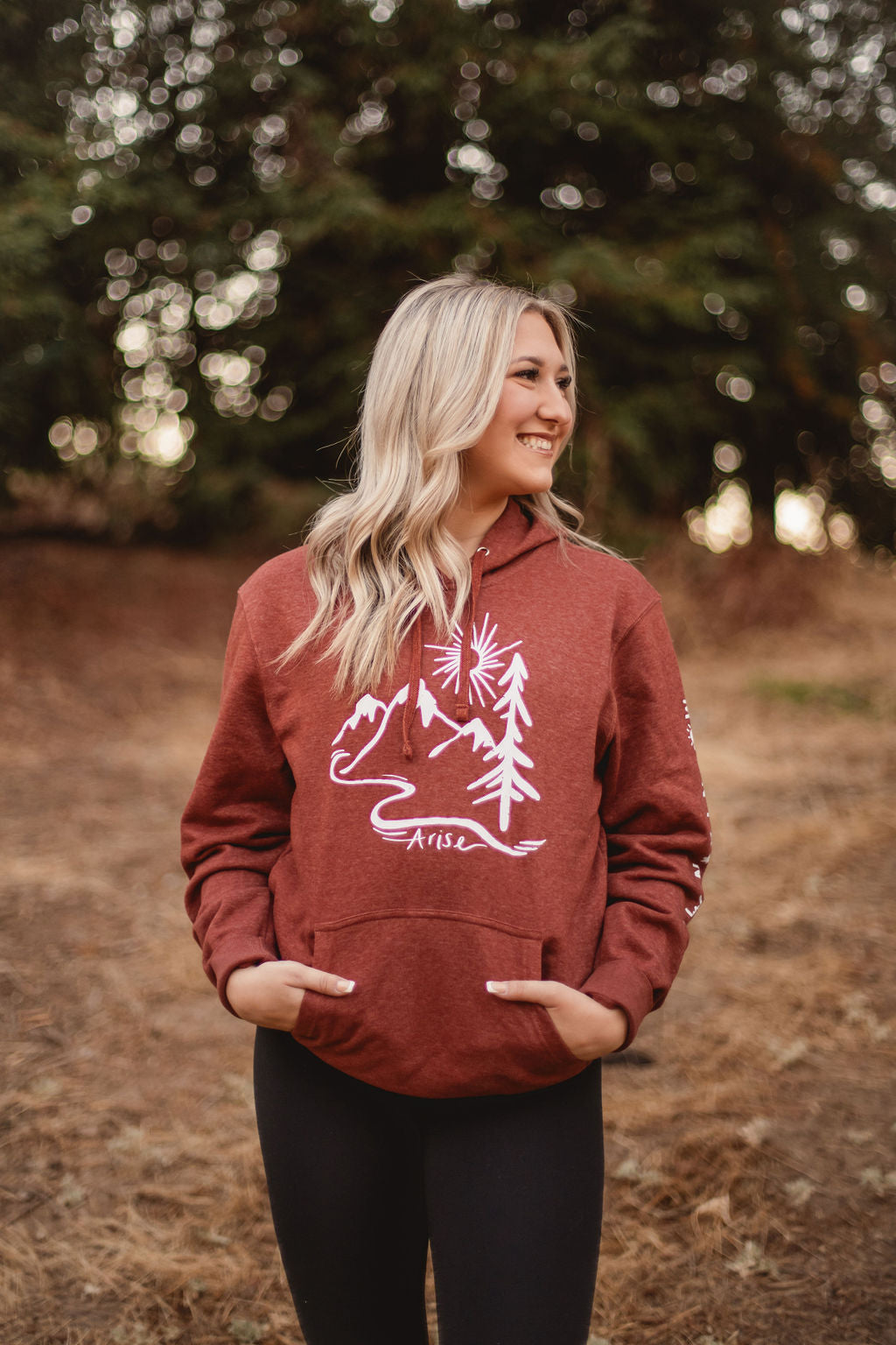 Woman wearing Rust colored Arise hoodie. Featuring white stamp with sun, mountain and tree