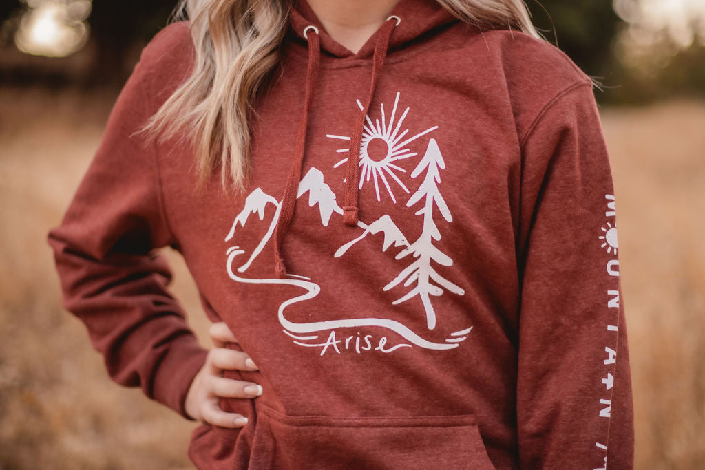 Rust colored Arise hoodie. Featuring white stamp with sun, mountain and tree