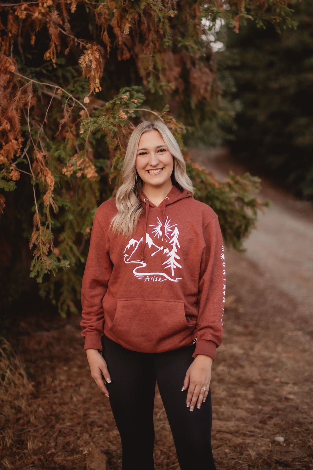 Woman wearing Rust colored Arise hoodie. Featuring white stamp with sun, mountain and tree