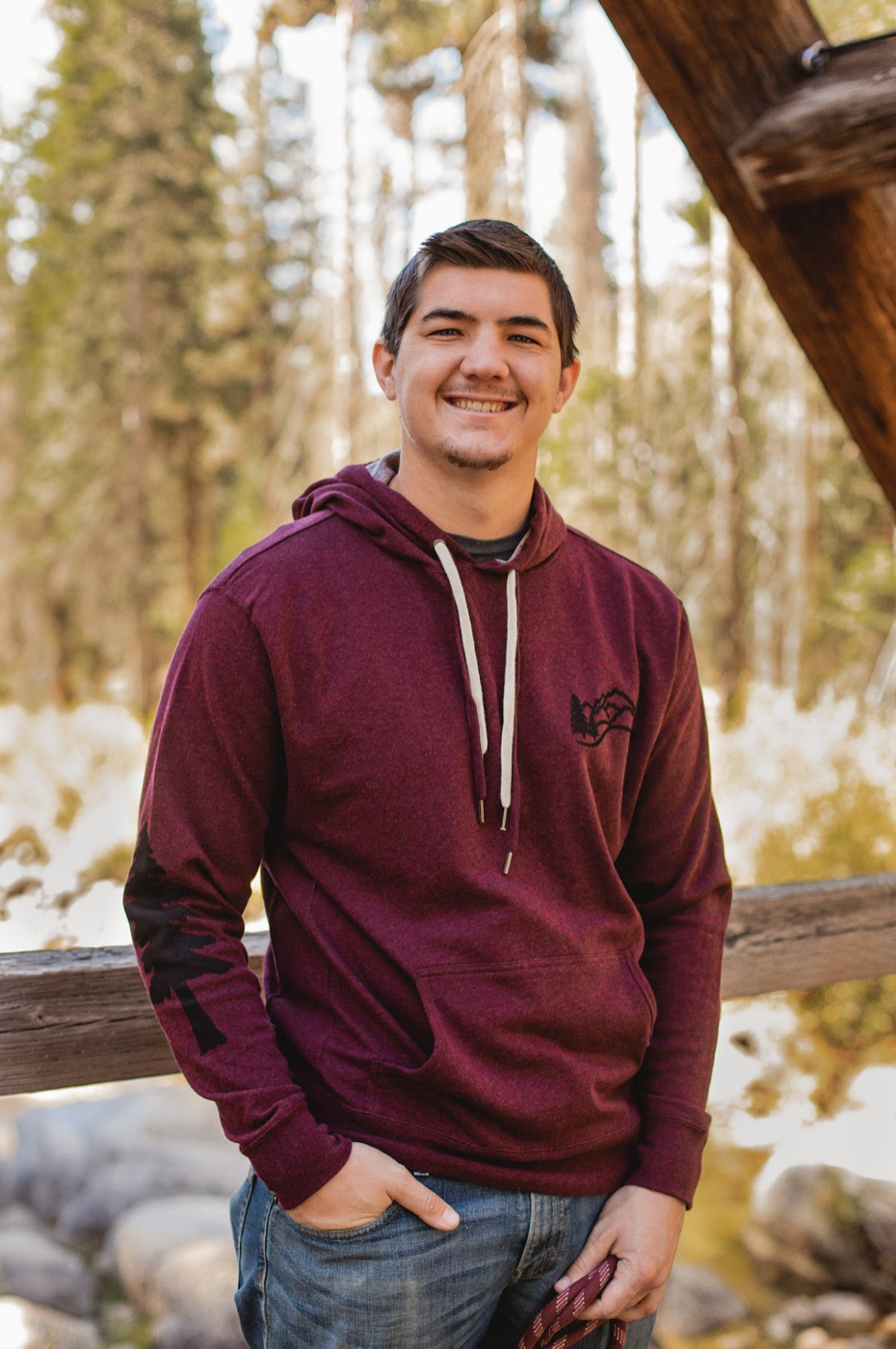 Man wearing maroon hoodie with tree stamp on right sleeve and tree with mountain scene on left shoulder